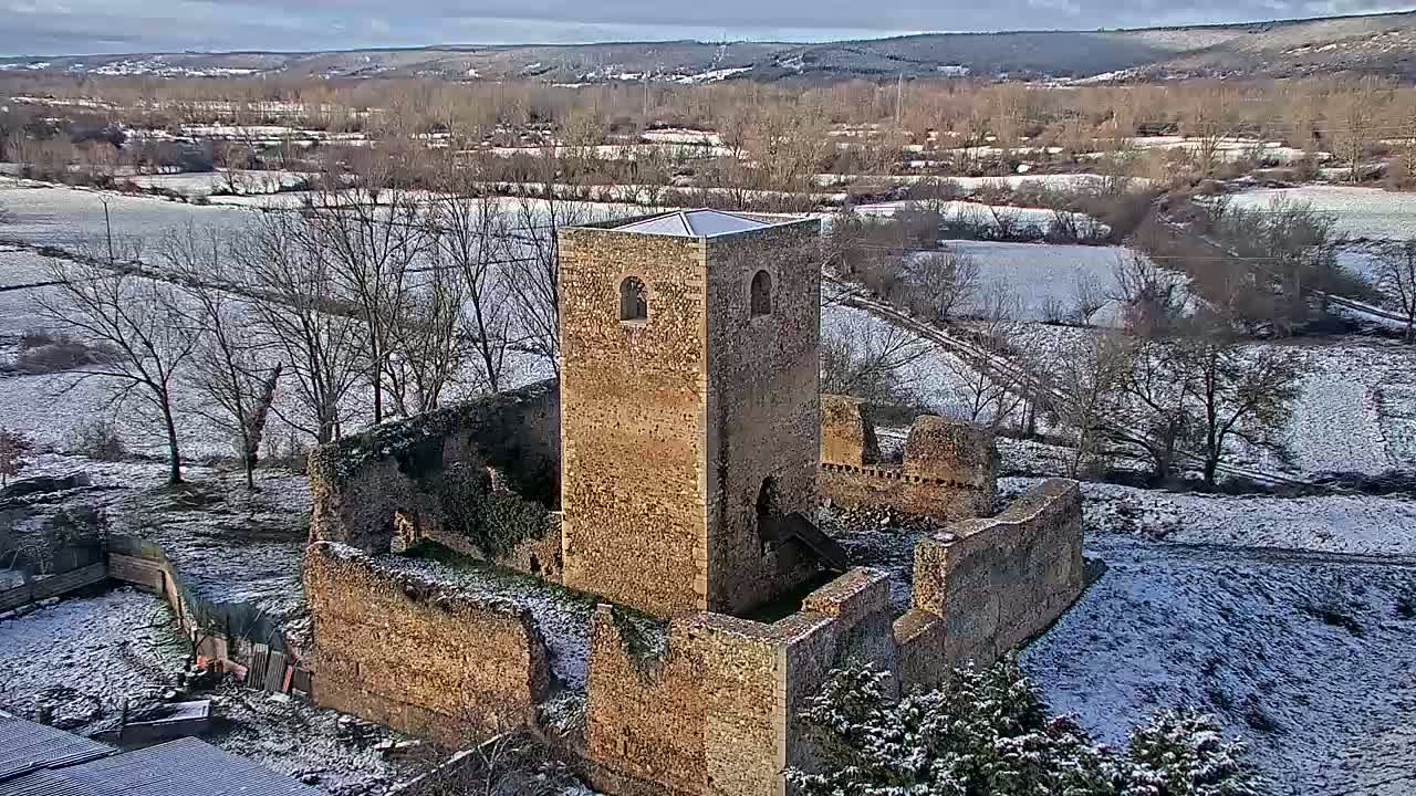 An aerial view shows the snow-covered ruins of a stone tower and surrounding buildings in a rural landscape.