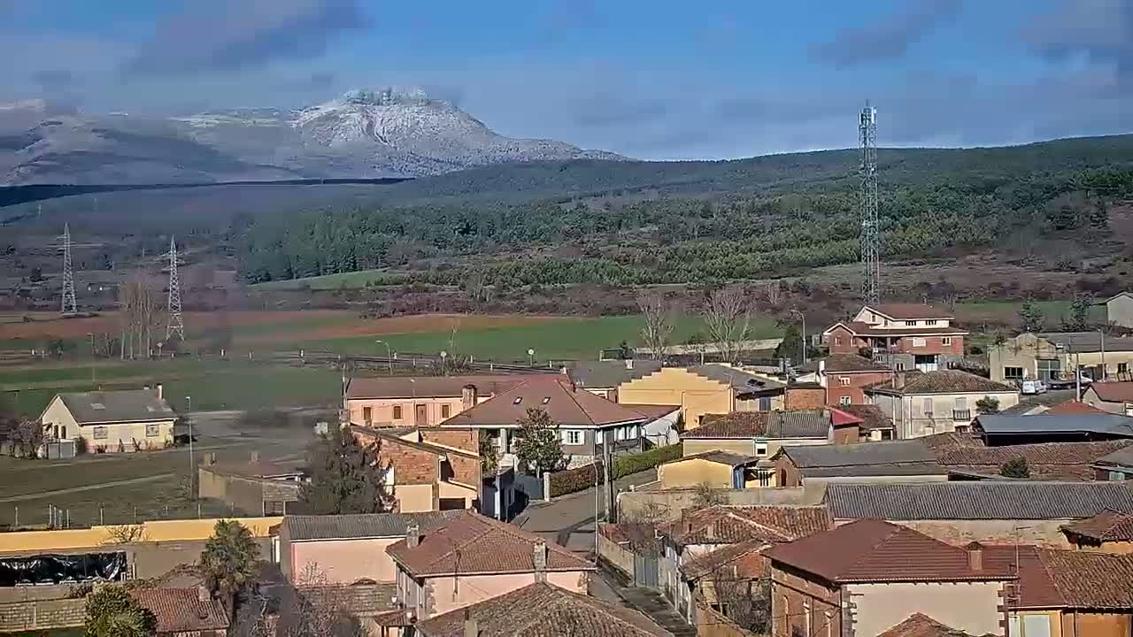 A small village with red-tiled roofs nestles in a valley below a snow-capped mountain on a clear, sunny day.