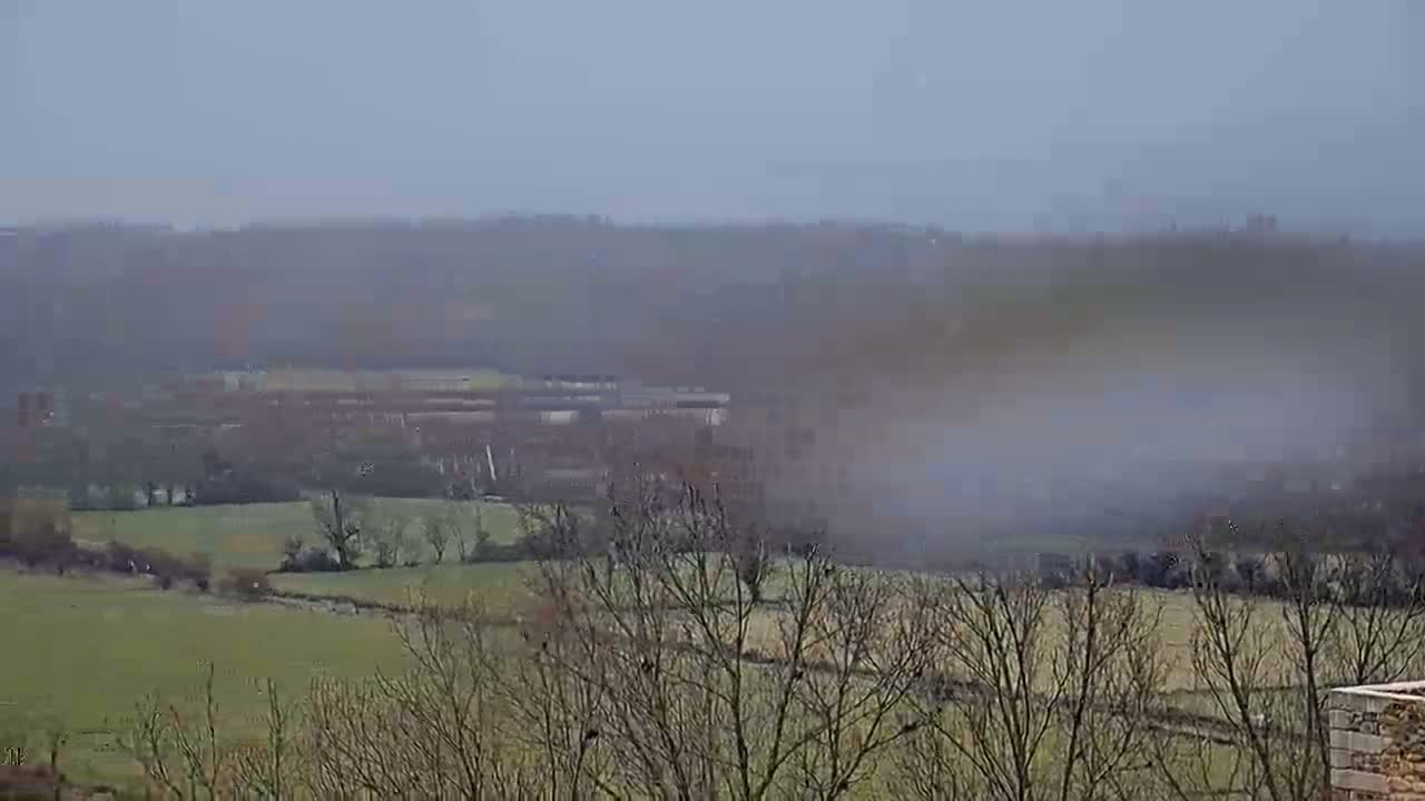 A hazy, overcast day reveals a rolling landscape of green fields and leafless trees, with a misty fog obscuring parts of the distant valley.