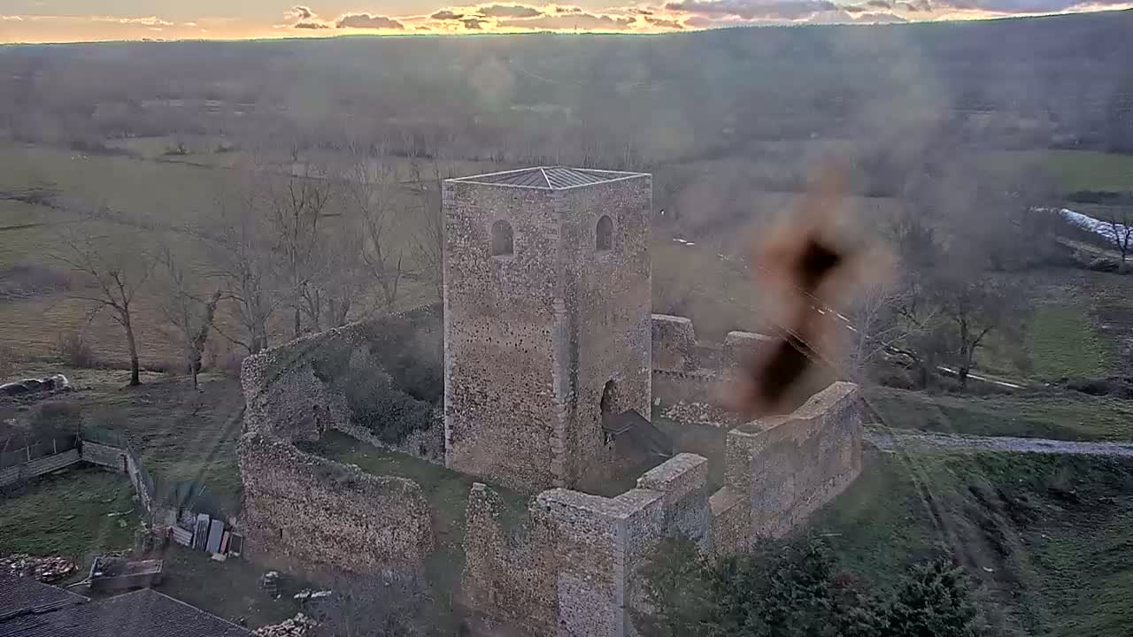 An aerial view shows the ruins of a stone tower and surrounding walls situated in a rural landscape under a partly cloudy sunset.