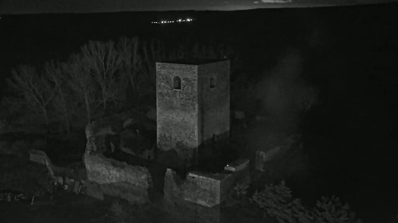 An aerial view at night shows the stone ruins of a tower and surrounding walls, partially obscured by trees under a dark sky.