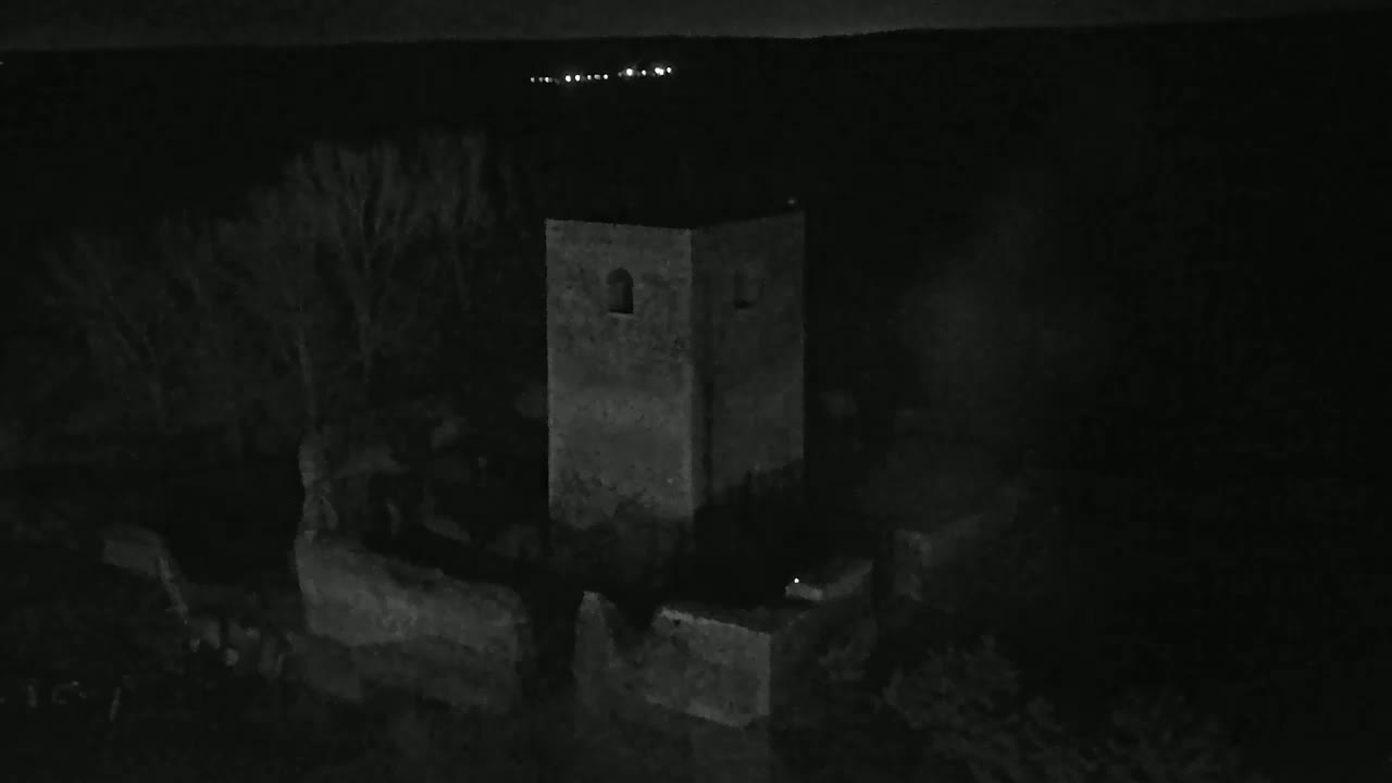 A stone tower stands in the darkness of night, partially obscured by trees and surrounded by low stone walls.
