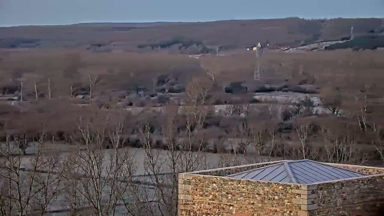 A stone structure with a metal roof sits in the foreground overlooking a flat, sparsely treed landscape under a dusky sky.