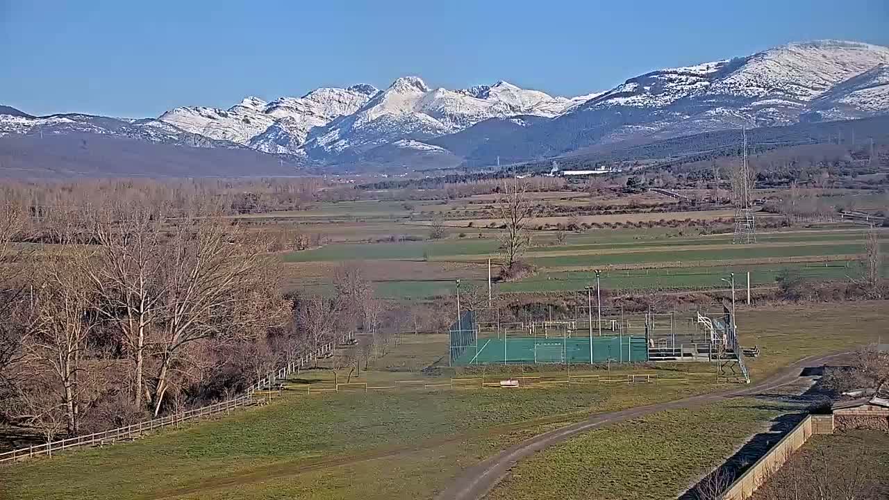A snow-capped mountain range is visible in the distance behind a flat, grassy field containing a sports court and leafless trees under a clear blue sky.