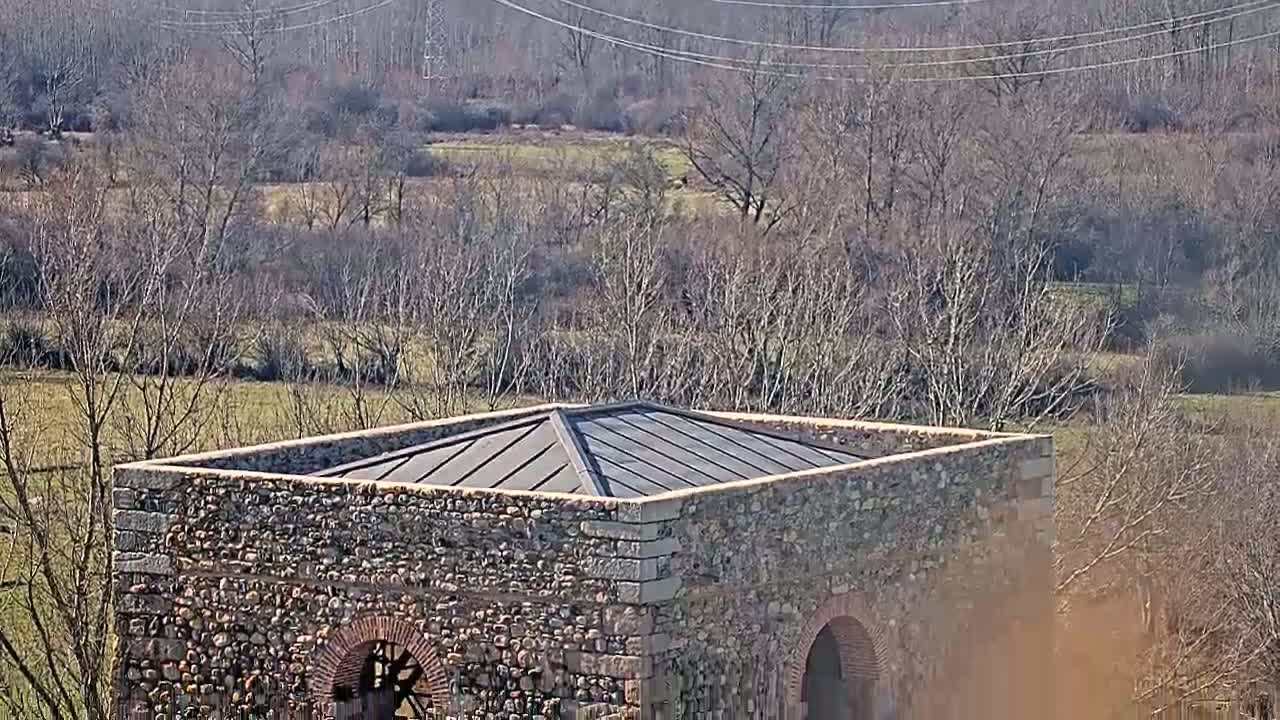 A stone structure with a flat, dark-colored roof sits in a field of bare trees under a sunny sky.