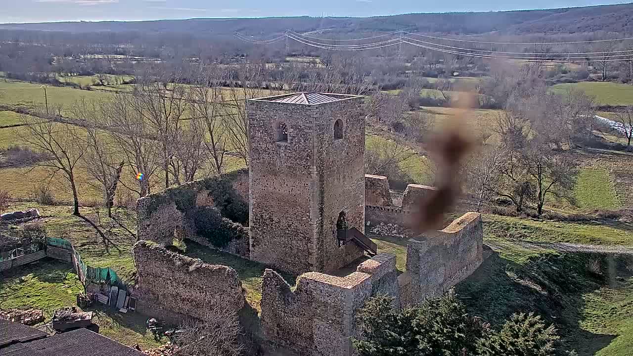 Stone ruins of a tower and surrounding walls are situated on a hillside overlooking a sunny, winter landscape of fields and bare trees.