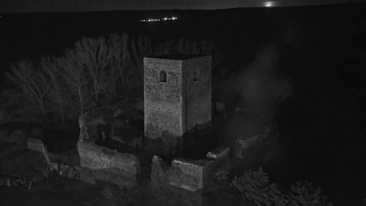 An aerial nighttime view shows stone ruins, including a tall standing tower, surrounded by bare trees under a dark sky.