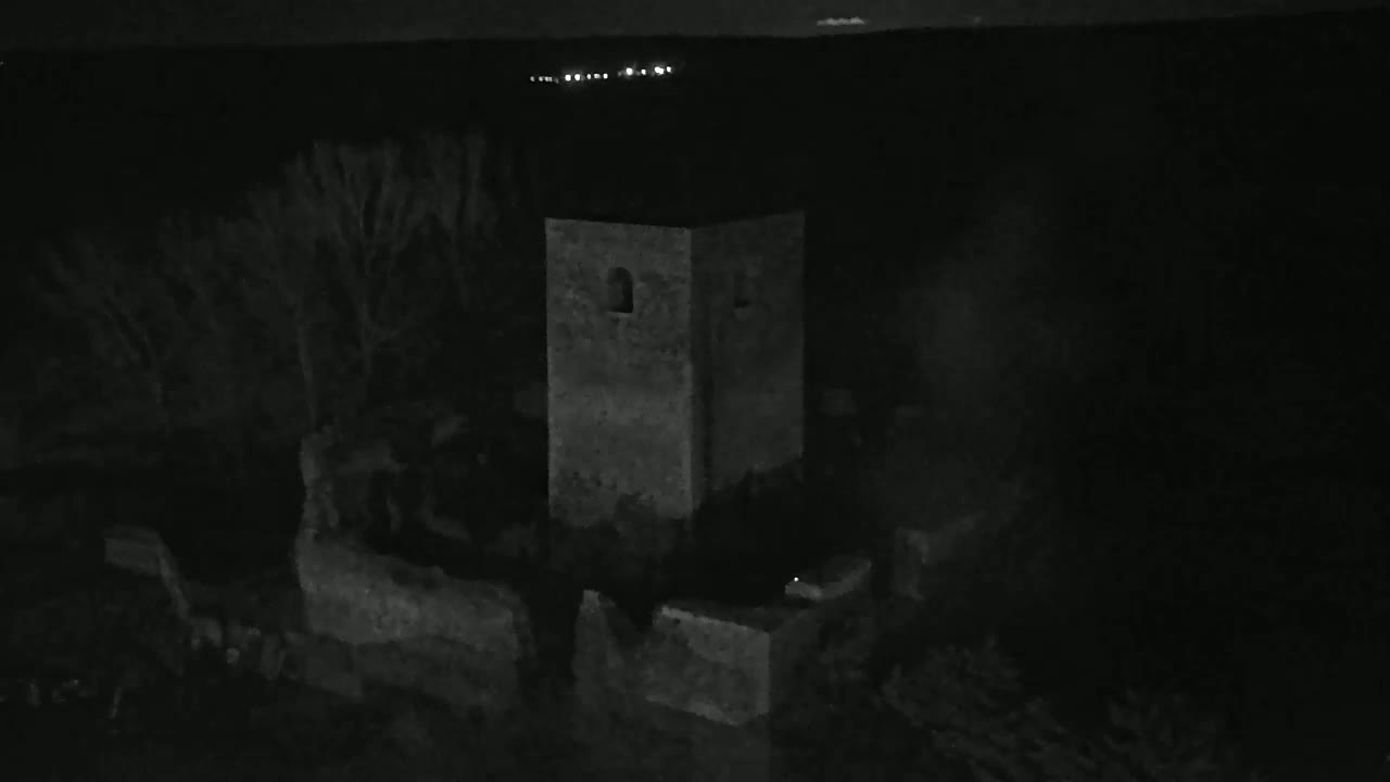 A stone tower stands in the darkness of night, surrounded by trees and low stone walls, under a seemingly clear sky with distant lights visible on the horizon.