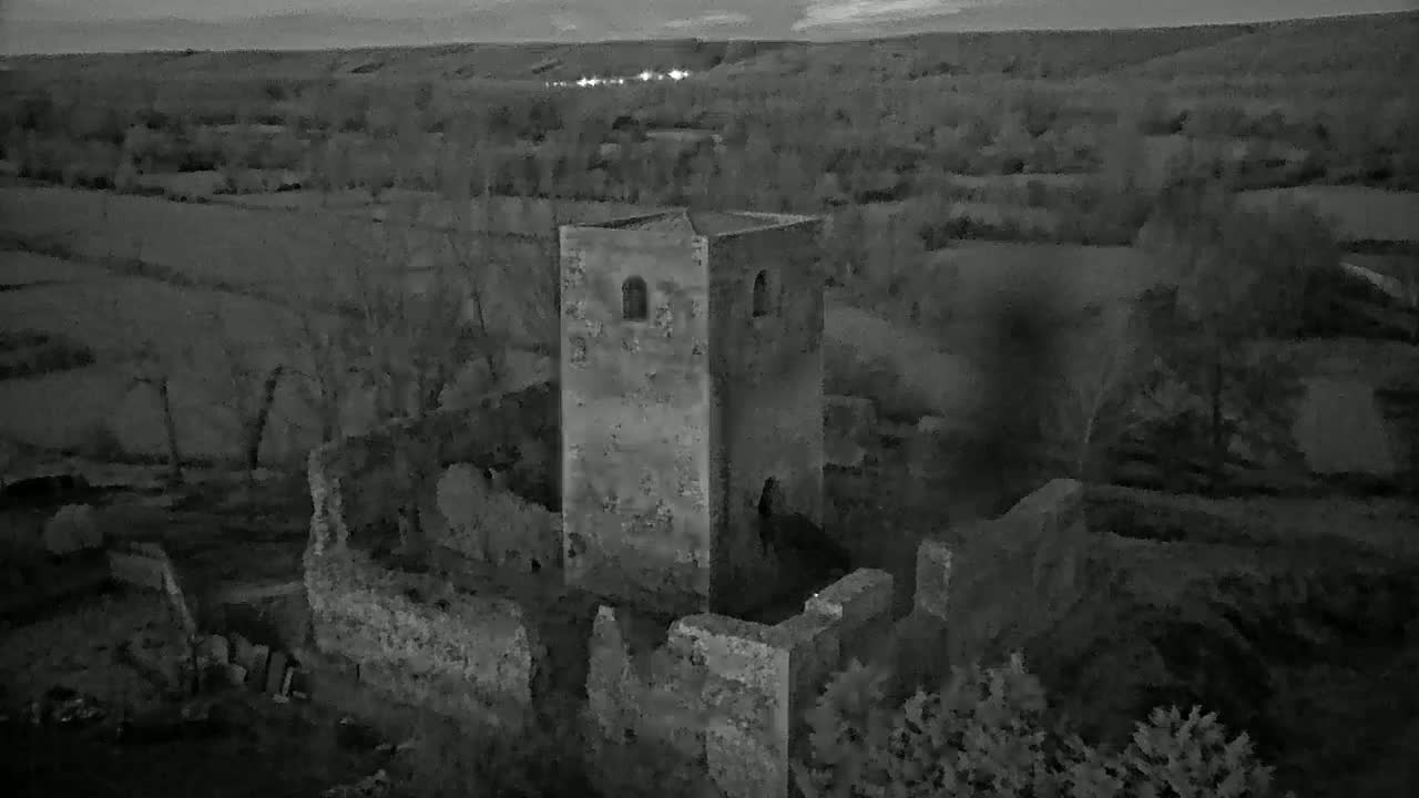 An aerial view shows the ruins of a stone tower and surrounding walls at twilight, overlooking a dark landscape.
