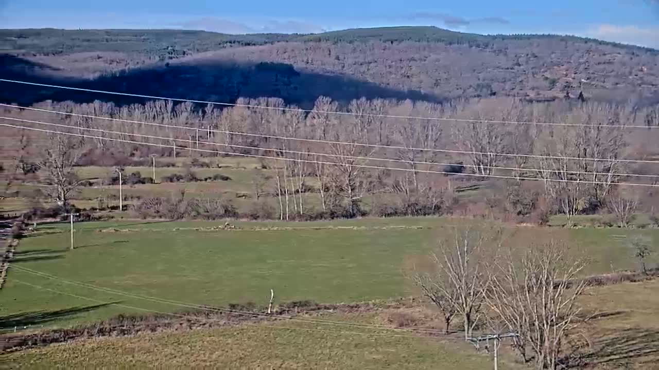 A partly sunny day reveals a rural landscape of fields, leafless trees, and power lines stretching across a valley, backed by distant hills.