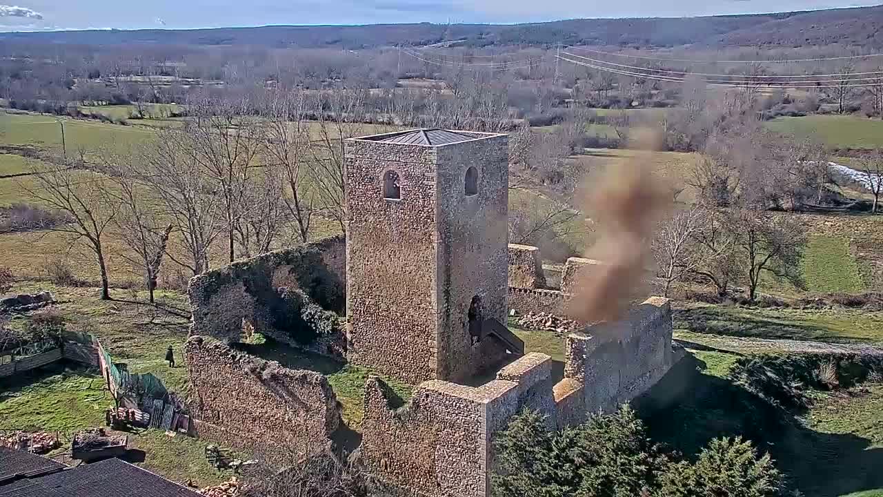 An aerial view shows the stone ruins of a castle or church on a sunny day, with brown fields and leafless trees in the surrounding countryside.