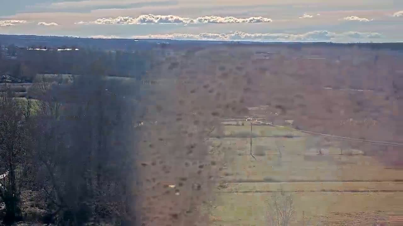 A long-range view of a mostly barren, flat landscape under a partly cloudy sky shows leafless trees and distant fields.
