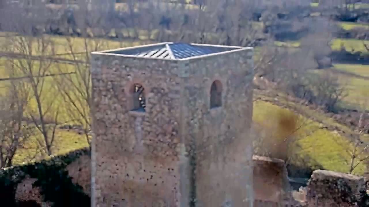 A stone tower with a flat roof sits amidst a landscape of bare trees and grassy fields under a bright, sunny sky.