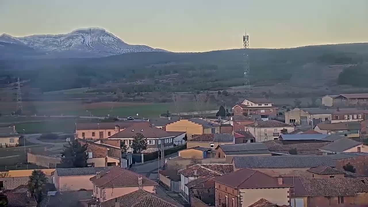A small town of red-tiled roofs nestles in a valley beneath a snow-capped mountain range under a clear, dusky sky.