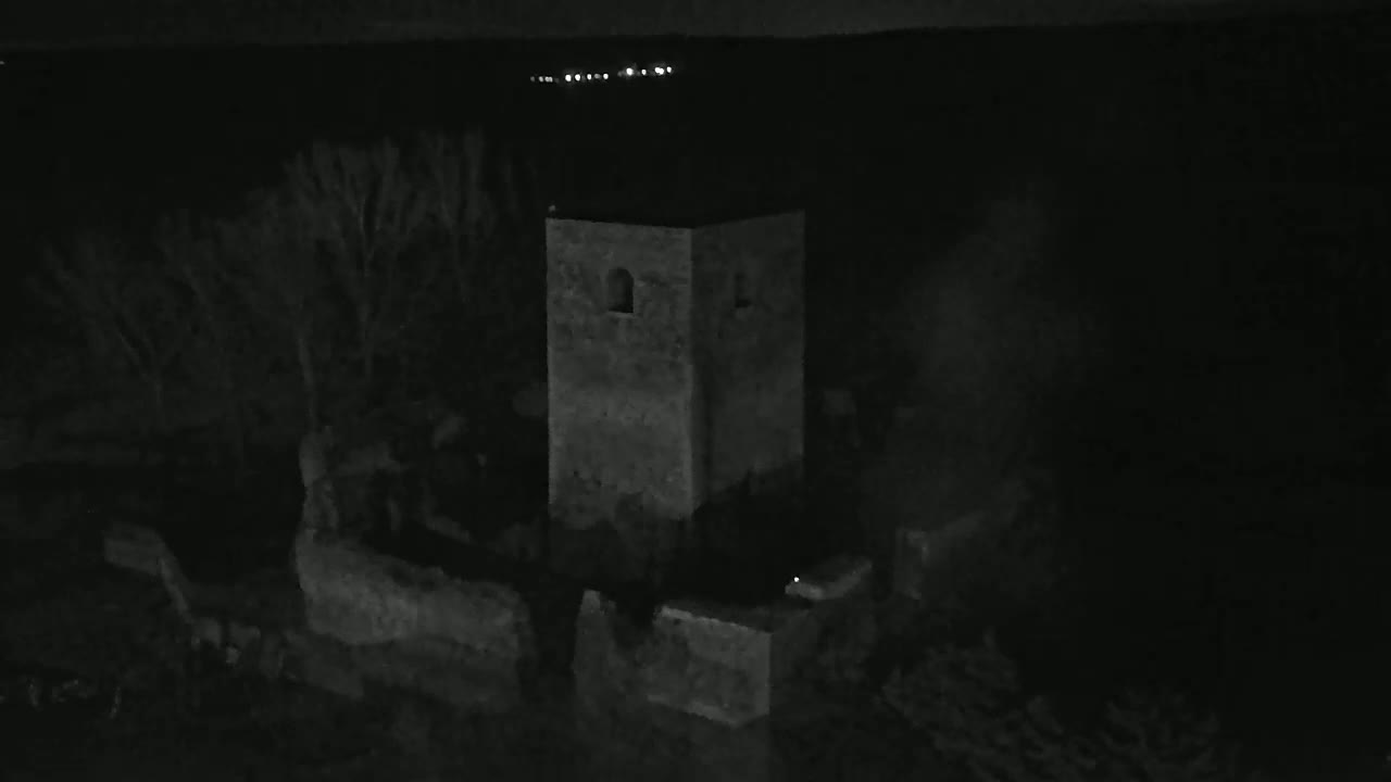 A stone tower and surrounding structures are visible at night, with a few distant lights in the background.