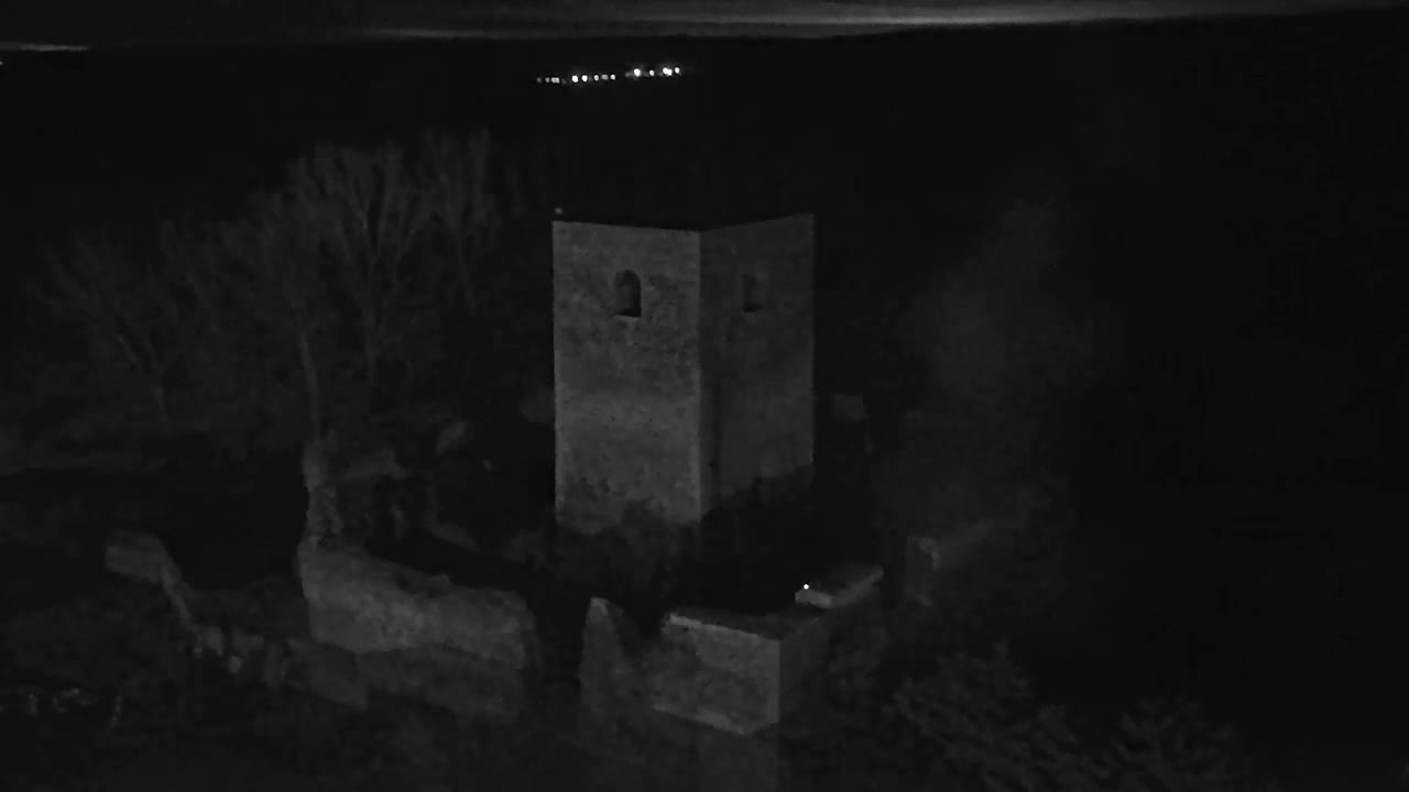 A stone tower and surrounding ruins are visible at night under a dark sky, with a few distant lights in the background.