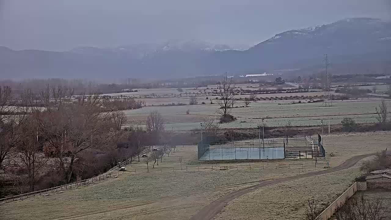 A frost-covered field with a paddle tennis court and bare trees stretches towards hazy mountains under a gray sky.