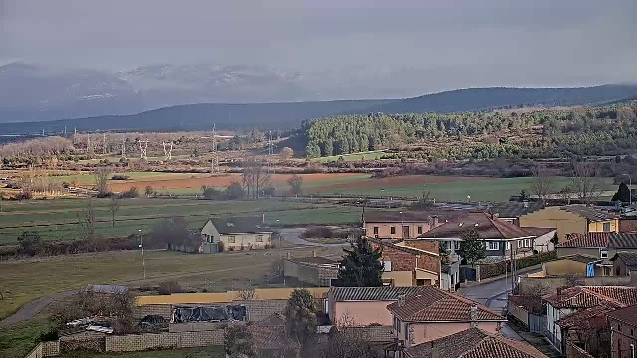 A small village with brown-tiled roofs sits in a valley, overlooking fields and a snow-capped mountain range under a mostly cloudy sky.