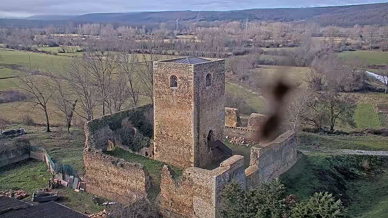 An aerial view shows the stone ruins of a tower and walls, set in a rural landscape under a cloudy sky.