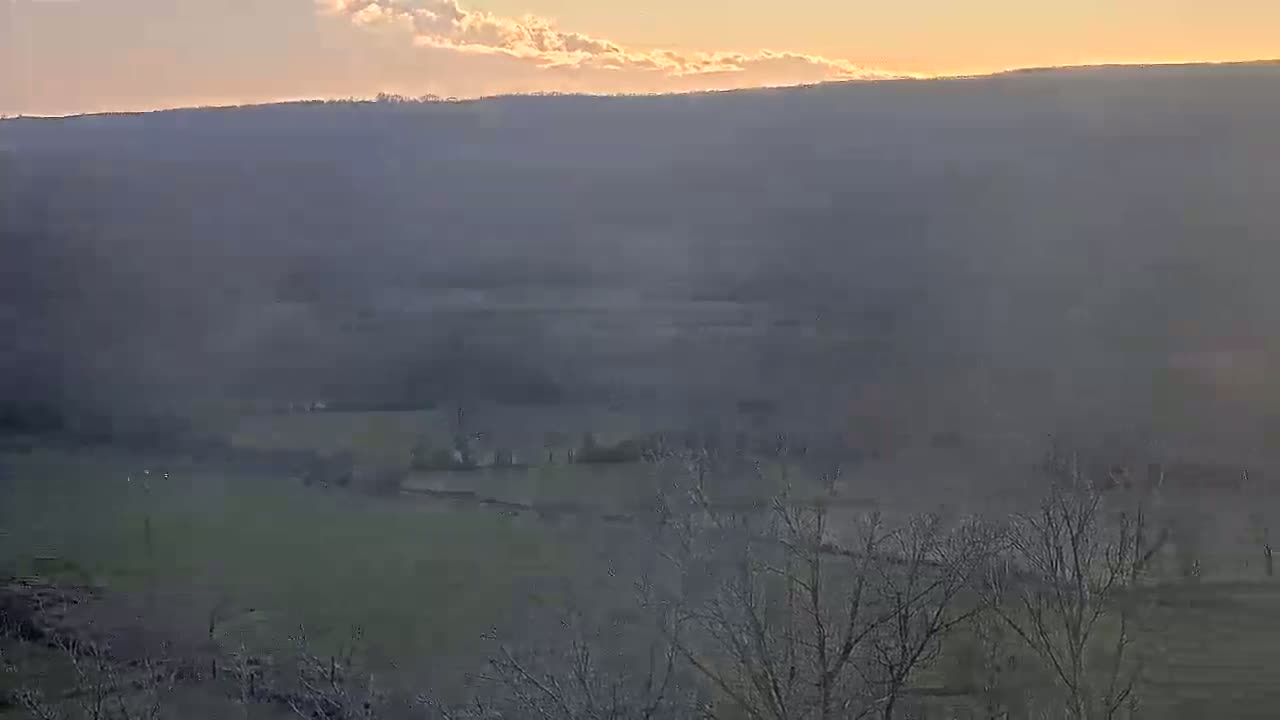 A hazy sunset illuminates a landscape of rolling hills, fields, and bare trees.