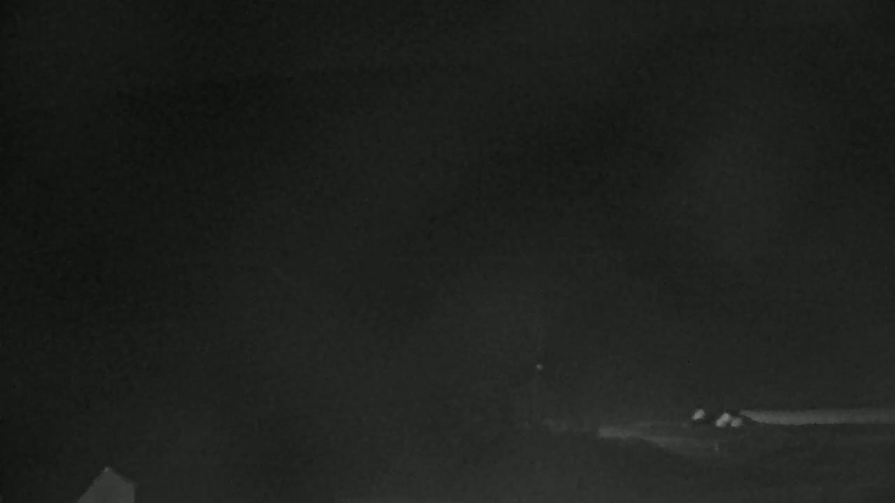 A dark, nighttime scene shows a few indistinct structures on a flat landscape under a mostly cloudy sky.