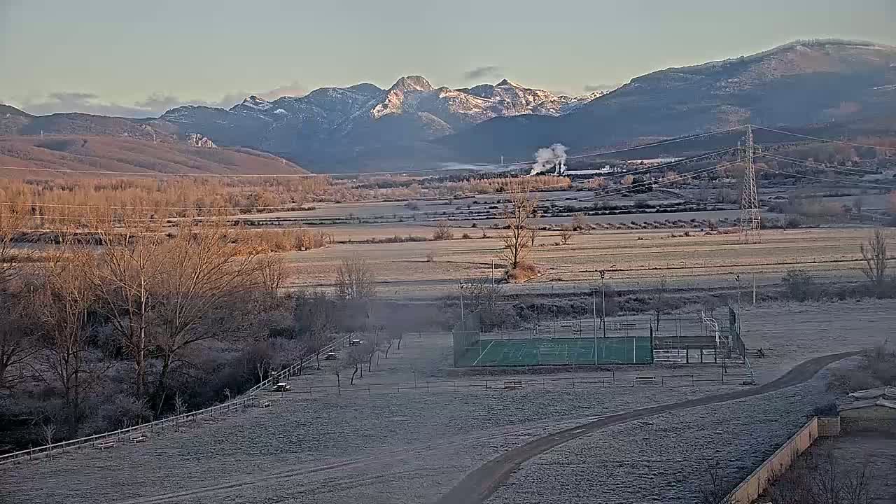 A frosty landscape shows a green sports court, barren trees, and distant snow-capped mountains under a clear sky.