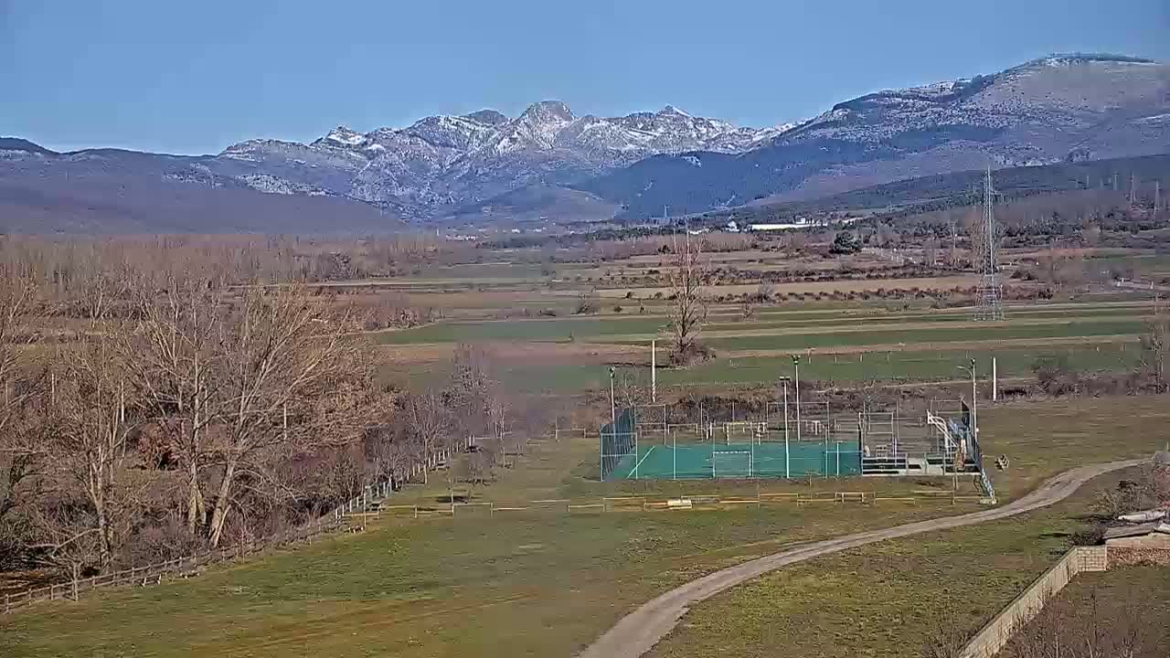 A green sports court is situated on a grassy field, overlooking a valley with leafless trees and snow-capped mountains under a clear blue sky.