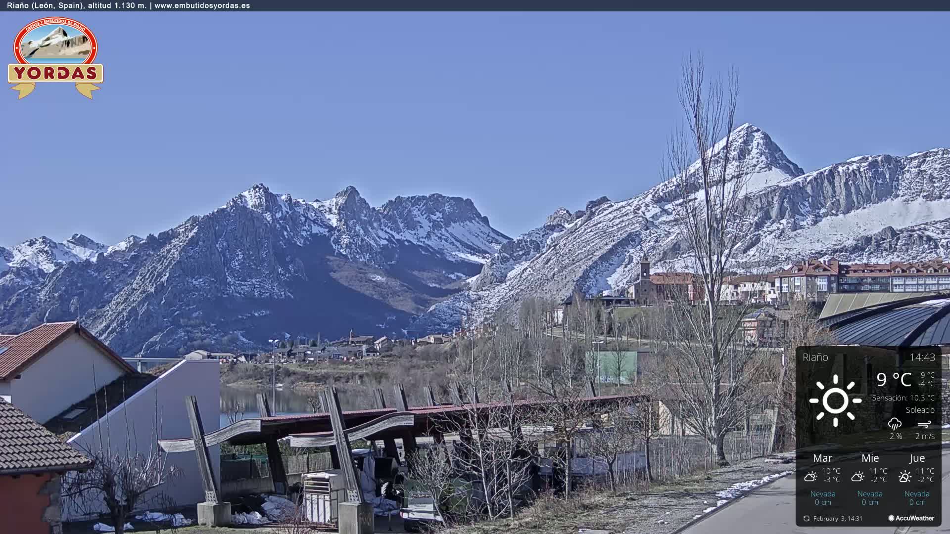 A sunny day reveals a snow-capped mountain range overlooking a town nestled beside a body of water, with a few buildings and a structure resembling a carport in the foreground.