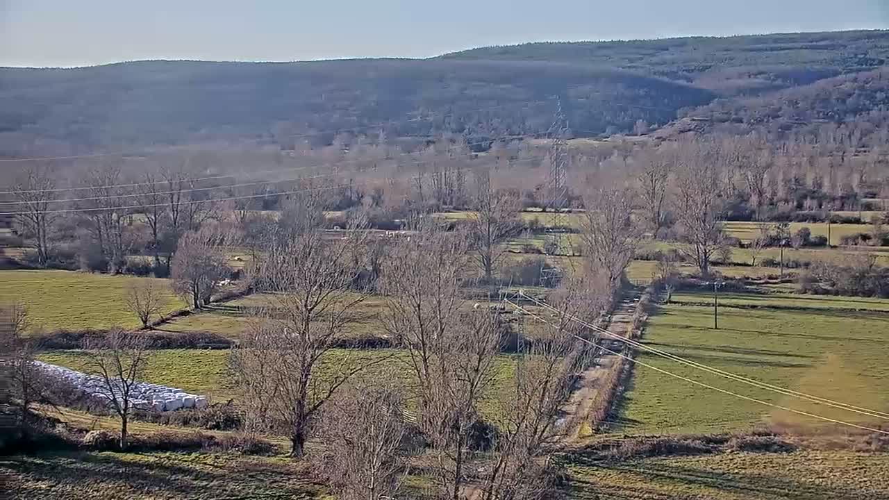 A high-angle view shows a sunny, rural landscape with leafless trees, grassy fields, a dirt road, and power lines stretching across the scene, with a range of hills in the background.