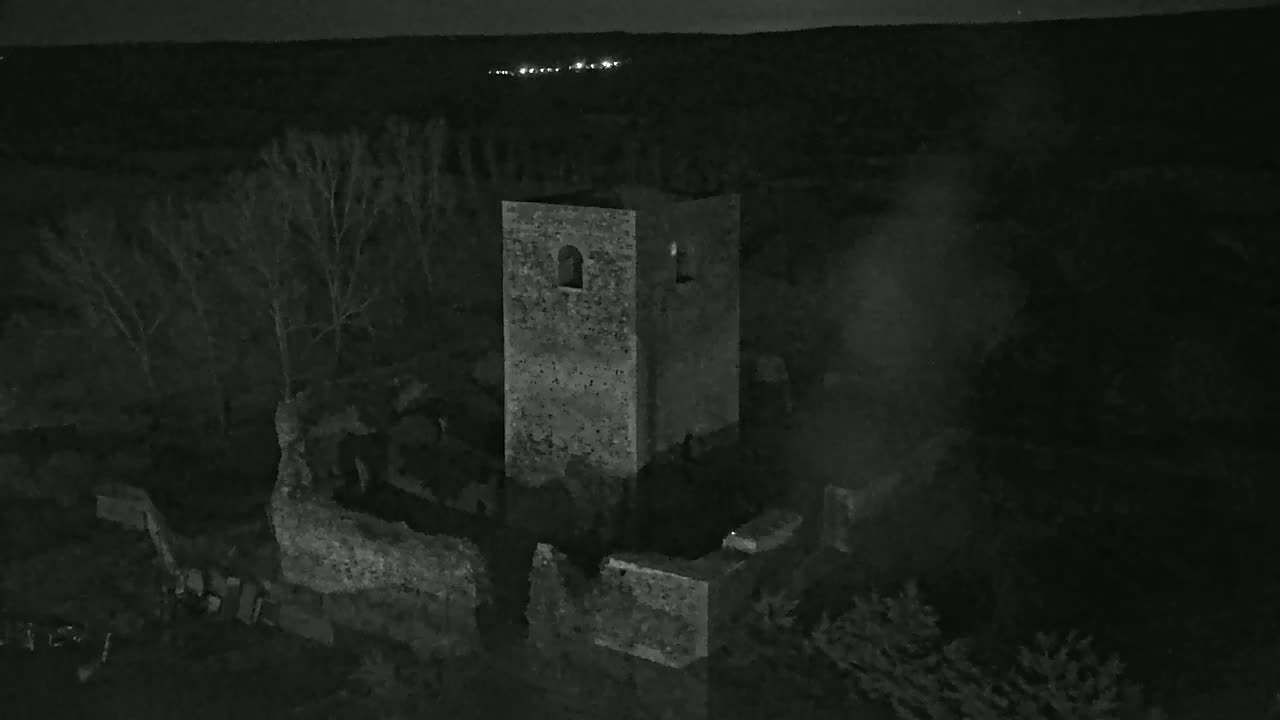 An aerial nighttime view shows stone ruins, including a standing tower, partially obscured by smoke and bare trees under a dark sky.