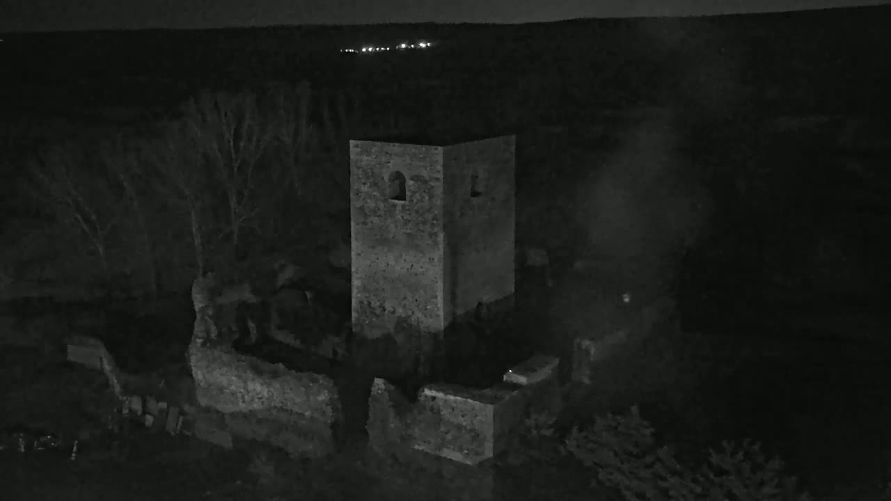 A stone tower, part of a larger stone ruin, is seen at night under a dark sky with distant lights visible on the horizon.