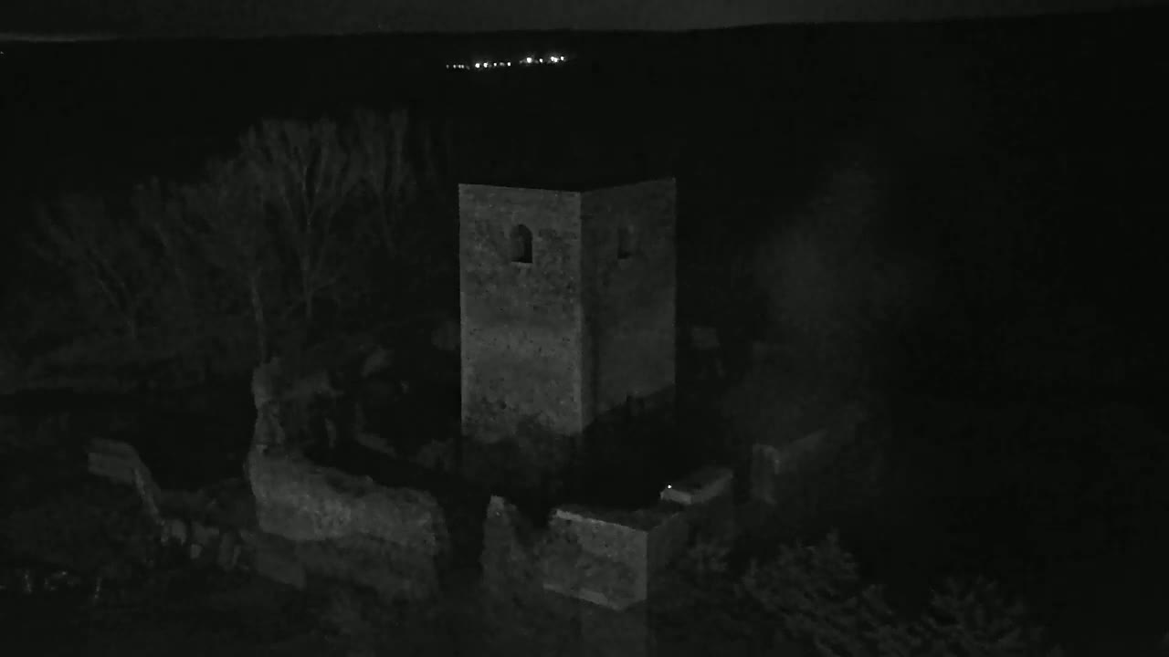 A stone tower, part of a larger ruin, stands silhouetted against a dark, night sky with distant lights visible in the background.