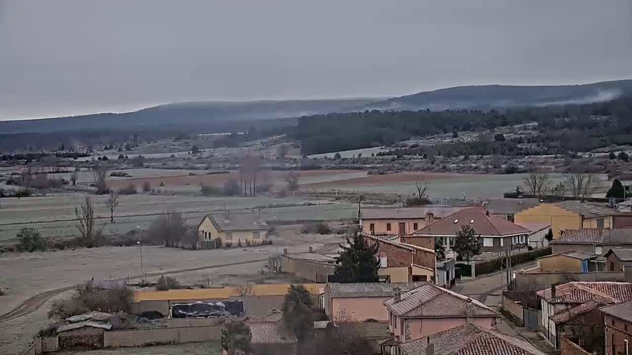 A frost-covered rural landscape with a small village of low-slung buildings nestled in a valley under a gray sky.