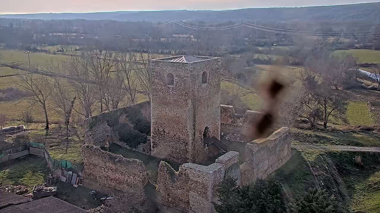 A partially ruined stone tower and surrounding walls sit in a rural landscape on a clear, sunny day.