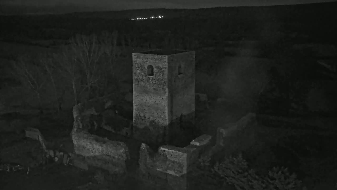 An aerial view at night shows the stone ruins of a tower and surrounding walls under a dark sky with distant lights.