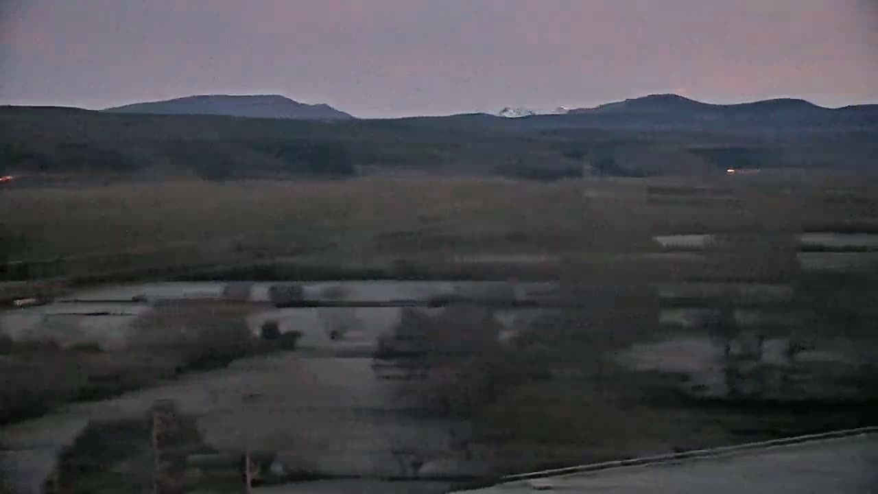 A dusky, low-light landscape shows a flat expanse of land with dark brown vegetation and a body of still water in the foreground, all under a pale, overcast sky, with distant dark hills visible on the horizon.