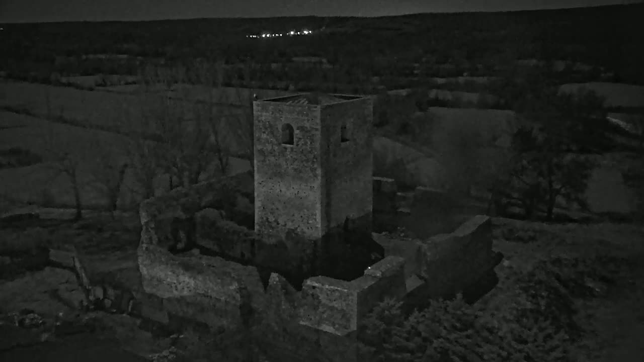 An aerial nighttime view shows the stone ruins of a tower and surrounding buildings amidst dark, sparsely vegetated land.