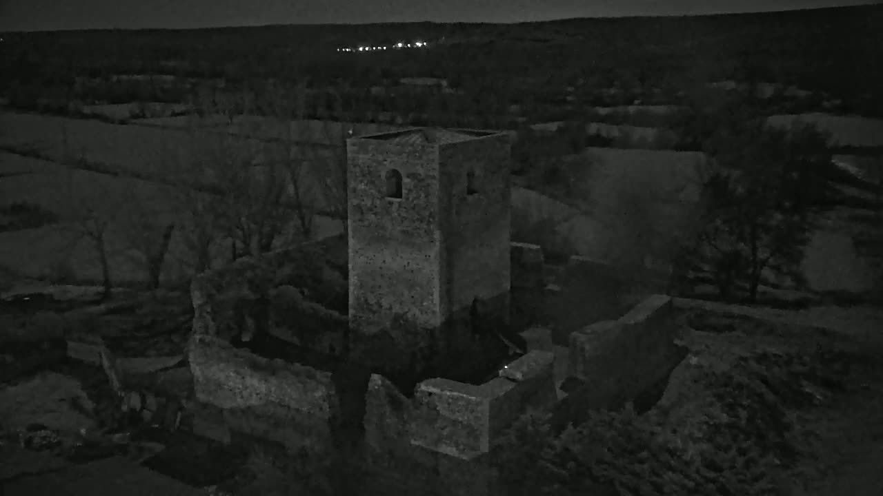An aerial nighttime view shows the ruins of a stone tower and surrounding walls in a dark, rural landscape.