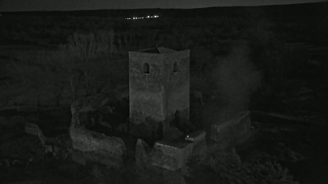 A stone tower, part of a larger ruined structure, stands at night under a dark sky, with distant lights visible on the horizon.