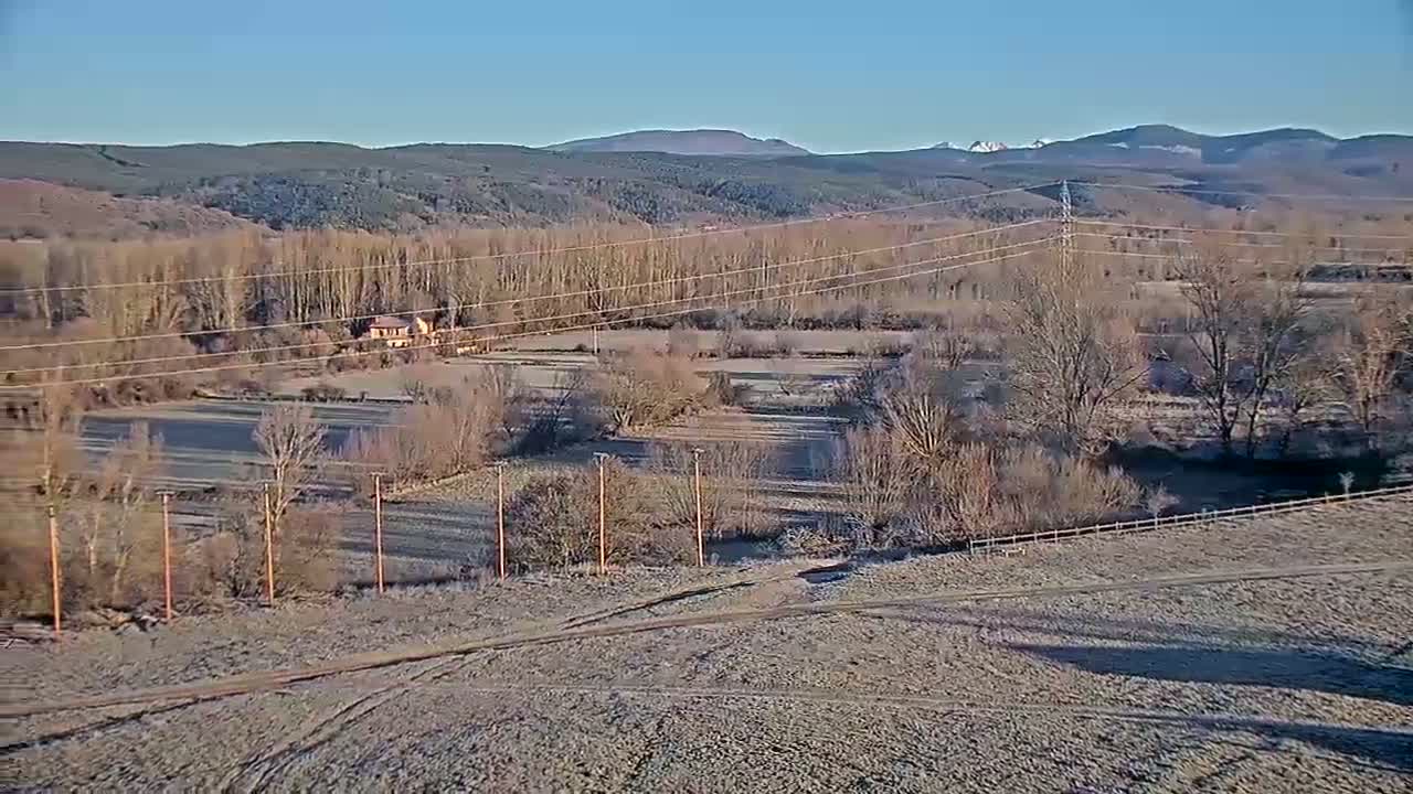 A frost-covered, rural landscape stretches out under a clear blue sky, featuring a line of trees, a small house, and power lines across a valley, with distant mountains visible in the background.