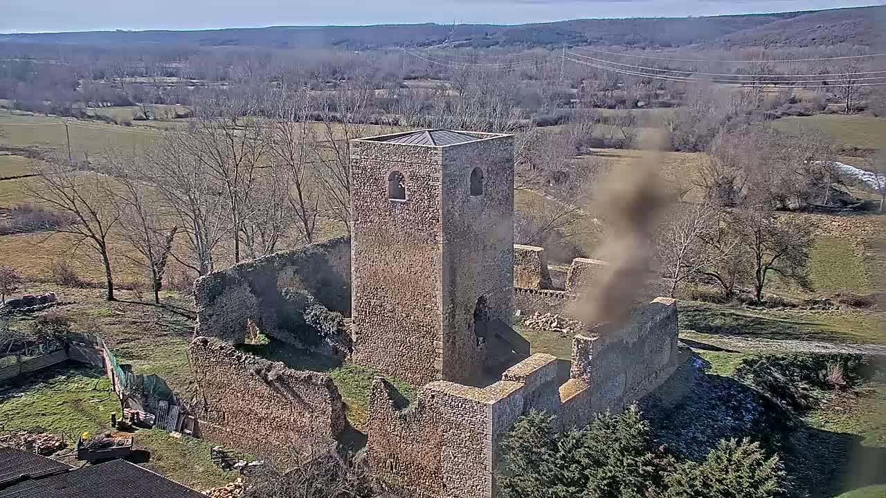 An aerial view shows the stone ruins of a building, possibly a church, with a standing tower, situated in a rural landscape under a clear, sunny sky.