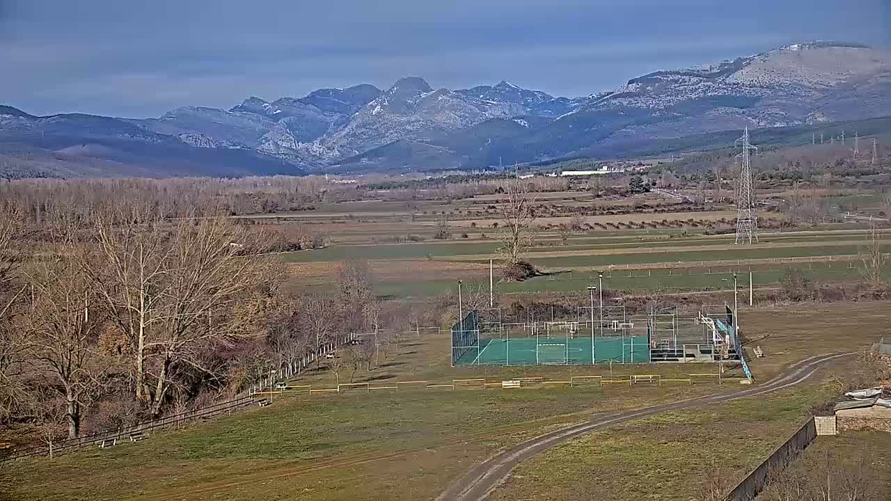 A paved outdoor court sits in a flat, grassy field, with leafless trees and snow-capped mountains in the background under a clear blue sky.