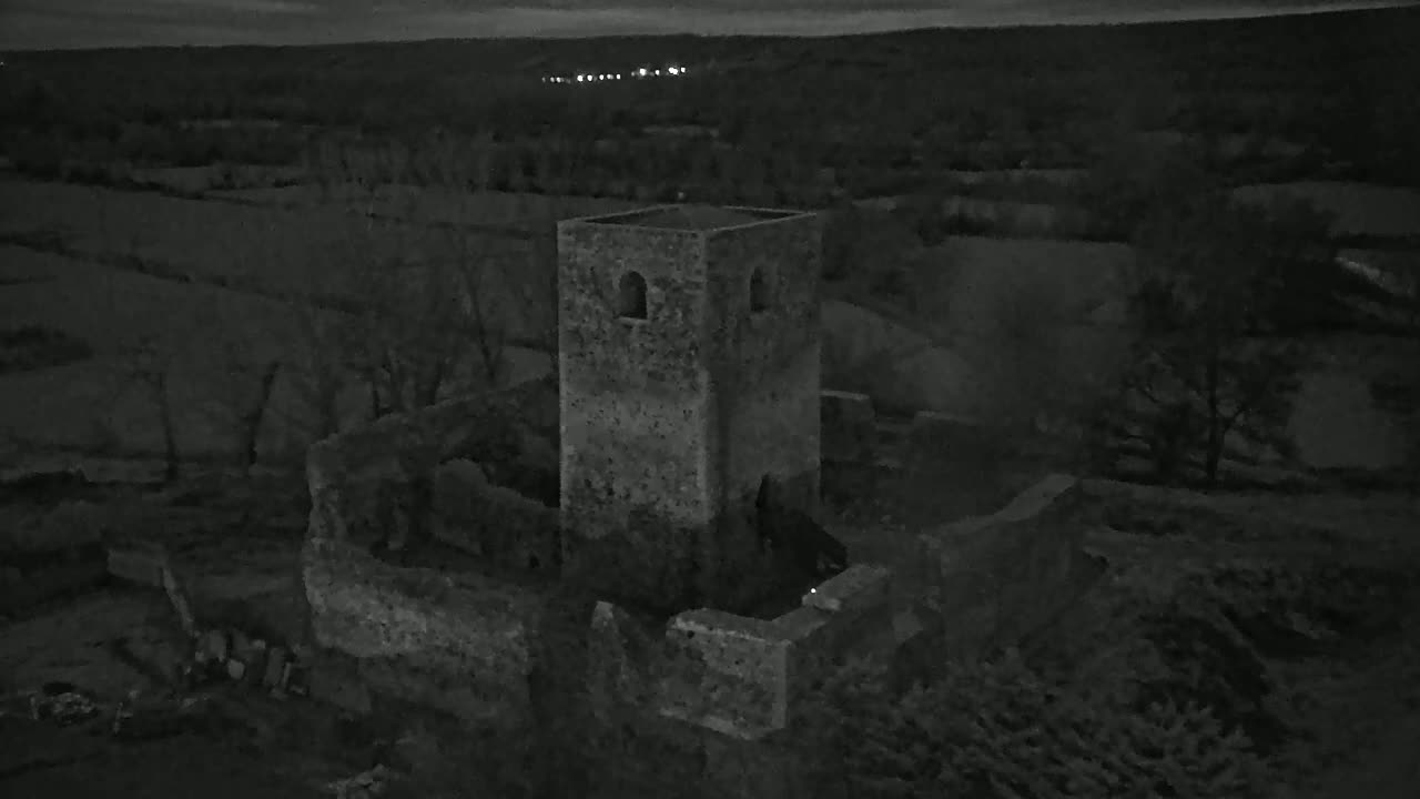 An aerial view at night shows the stone ruins of a tower and surrounding buildings in a rural landscape under a dark sky.