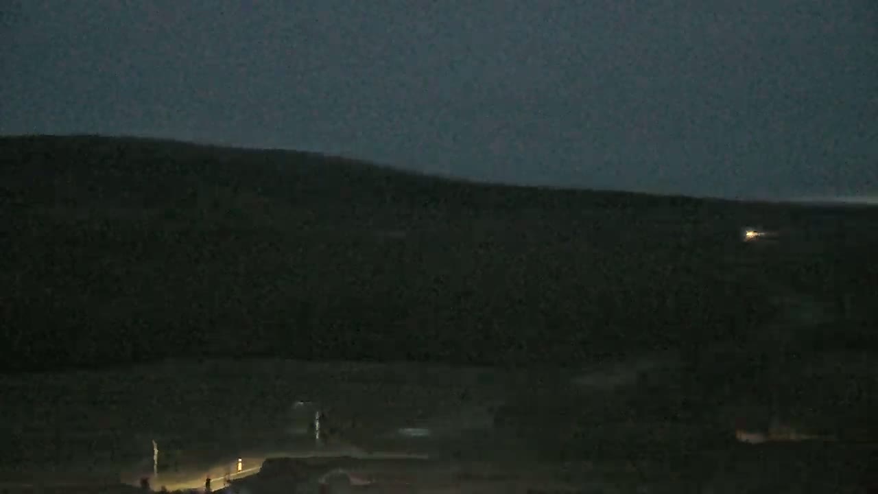 A dark, nighttime landscape shows a distant hill under a dusky sky, with a few faint lights visible in the lower portion of the frame.