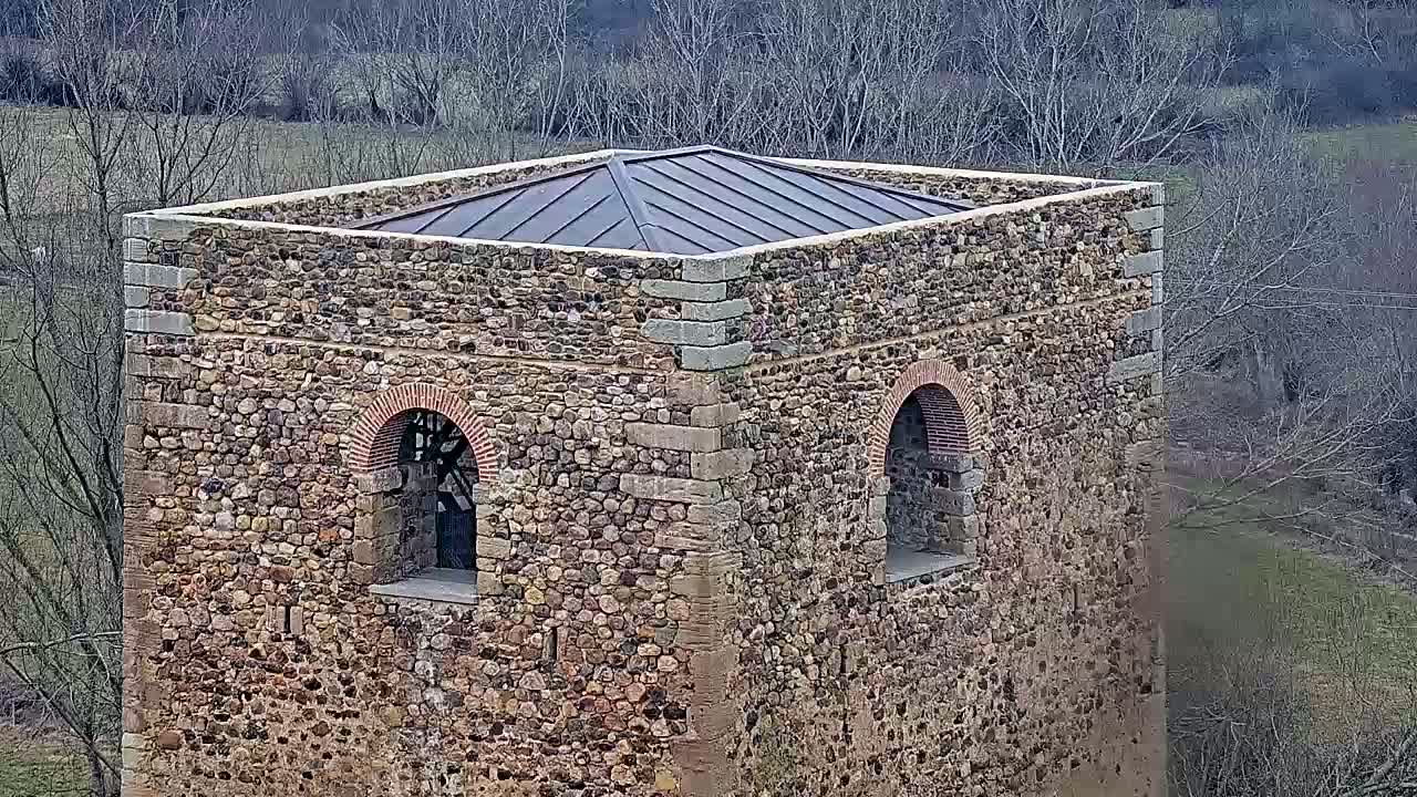 A stone tower with a dark metal roof stands overlooking a landscape of bare winter trees and a grassy field.