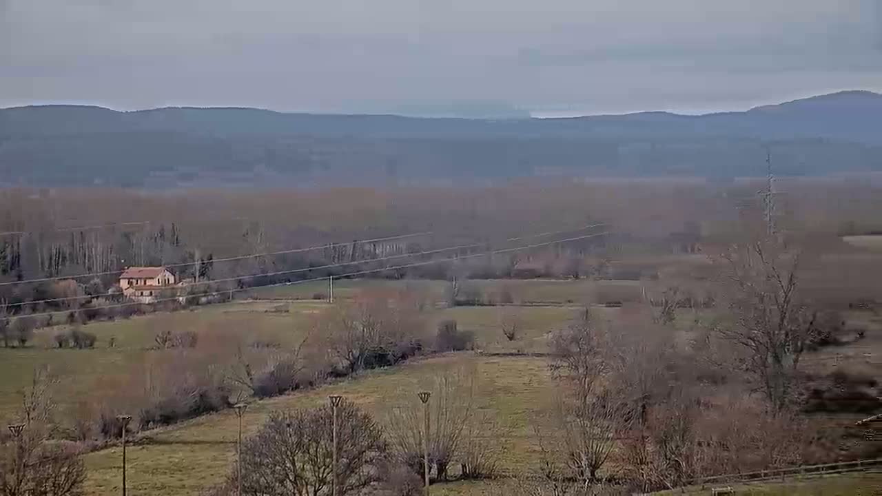 A pale, overcast sky hangs over a flat, grassy landscape dotted with leafless trees and a lone house, with distant, hazy mountains in the background.