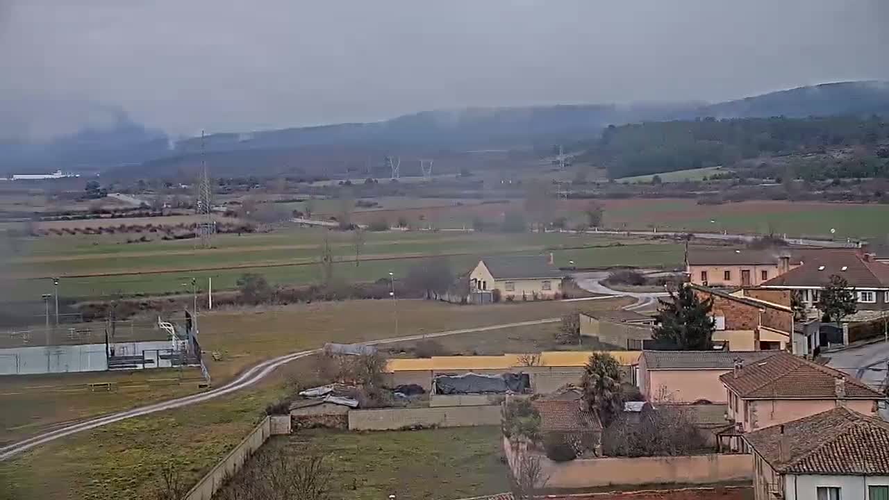 A hazy, overcast day reveals a rural landscape with farmlands, a small village of low-slung buildings, and a distant line of hills.