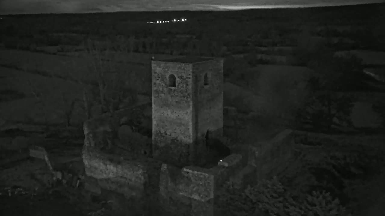An aerial view at night shows the stone ruins of a tower, surrounded by dark trees and fields under a dusky sky.