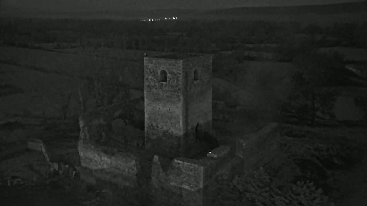 An aerial nighttime view shows the stone ruins of a tower and surrounding walls in a dark, rural landscape.
