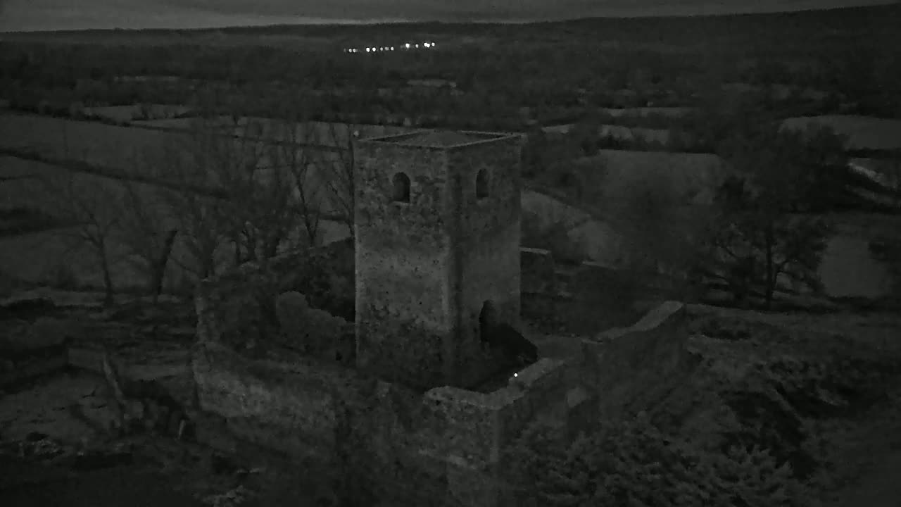 An aerial view at night shows the stone ruins of a tower and surrounding buildings amidst a dark, sparsely wooded landscape.